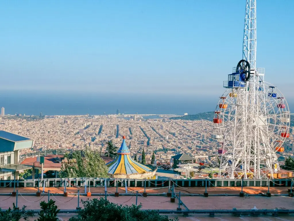 Vistas de Barcelona desde el mirador de la montaña del Tibidabo. En primer plano, aparece una noria de colores, la atalaya y un carrusel.